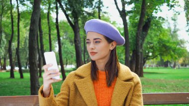 Şık sonbahar kıyafetleri içinde genç bir kadın parkta cep telefonuyla mesajlaşıyor.