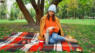 Parkta piknik yaparken termos açan şık sonbahar elbiseli bir kadın.
