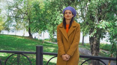 Güz parkında çitlerin yanında duran sonbahar kıyafetli moda kadın.