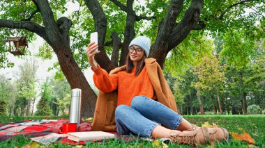 Parkta piknik yaparken cep telefonuyla selfie çeken şık sonbahar kıyafetli bir kadın.