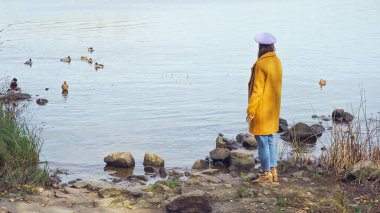 Güz kıyafetli bir kadın gölün kenarında duruyor parkta ördekler var.