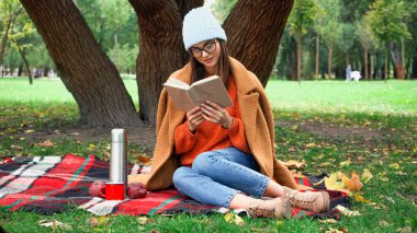 Sonbahar kıyafetli genç bir kadın parkta piknik yaparken kitap okuyor.