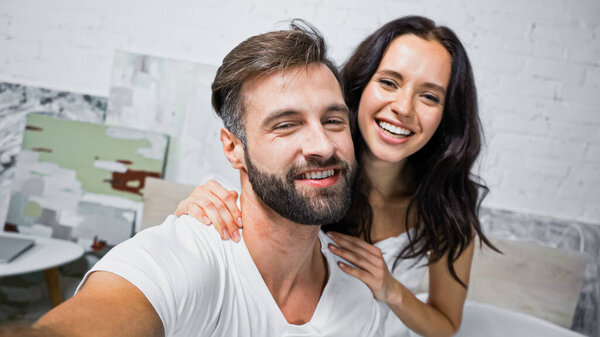 happy bearded man and brunette woman smiling at camera in bedroom