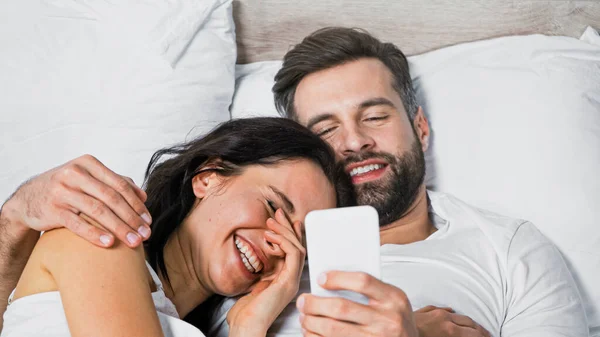 smiling man using mobile phone while hugging excited girlfriend in bed