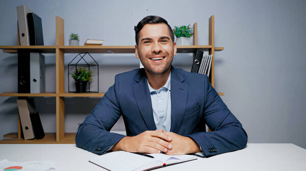 Smiling businessman looking at camera near notebook and papers on blurred foreground 