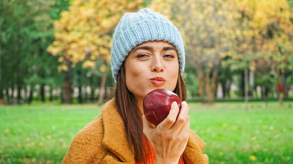 joyful woman looking at camera while eating juicy apple in park