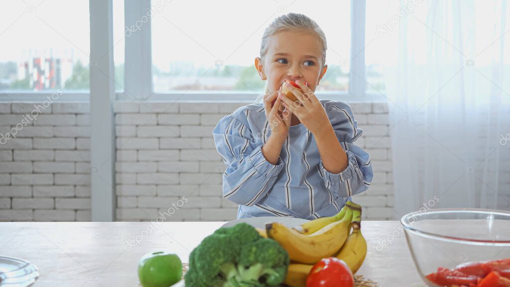 Girl eating baguette near fresh vegetables and fruits on blurred foreground 