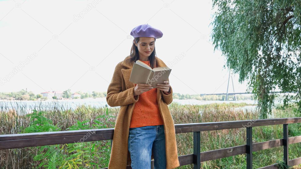 fashionable woman reading book while standing on bridge in autumn park