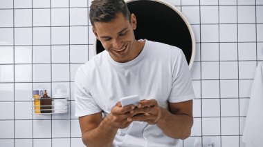 Beyaz tişörtlü gülümseyen adam banyoda akıllı telefondan sohbet ediyor.