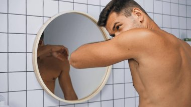 shirtless man smelling himself near mirror in bathroom clipart