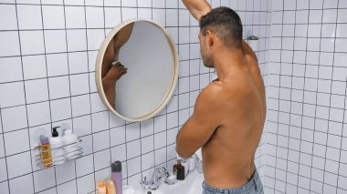 Üstsüz adam banyoda aynanın yanına deodorant sürüyor.