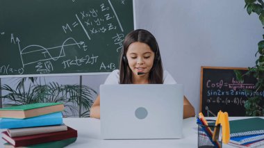 Evde internetten ders çalışırken bilgisayarına bakan kulaklıklı mutlu okul çocuğu.