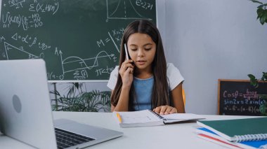 Liseli kız akıllı telefondan konuşuyor ve masadaki dizüstü bilgisayarın yanına bakıyor.