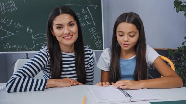 mother smiling near daughter doing homework at home clipart