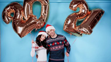 Noel Baba şapkalı genç çift mavi arka planda 2021 işareti şeklinde balonlar tutarken sarılıyorlar.