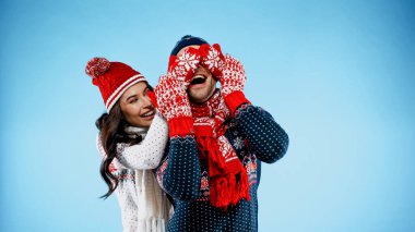 Cheerful woman in sweater and mittens covering eyes of boyfriend on blue background clipart
