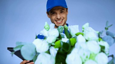 happy delivery man holding fresh flowers on blurred foreground isolated on blue clipart