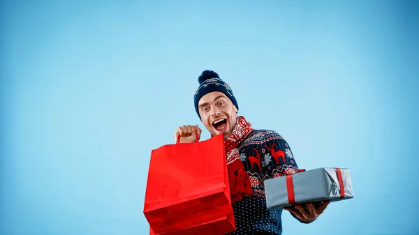 Excited man in sweater holding shopping bags and present on blue background