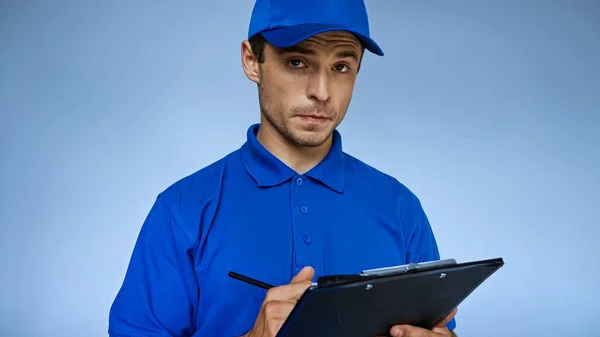 serious delivery man writing order on clipboard isolated on blue