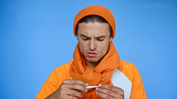 Hombre Enfermo Disgustado Gorro Caliente Bufanda Mirando Termómetro Aislado Azul — Foto de Stock