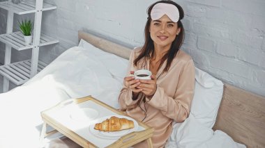 happy woman holding cup of coffee near croissant on tray in bed clipart