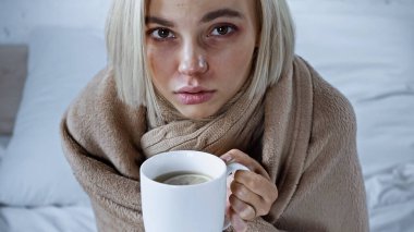 sick woman, wrapped in blanket, holding cup of warm tea in bedroom