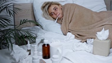 Hasta kadın yatakta yatarken sıcaklığını ölçüyor. Ön planda bulanık ilaçlar var.