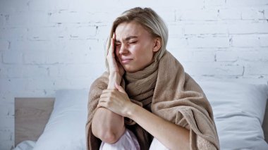 ill woman, wrapped in warm blanket, suffering from headache in bedroom