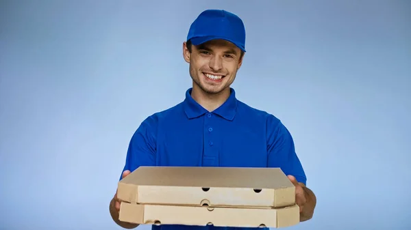 happy delivery man holding pizza boxes while smiling at camera isolated on blue