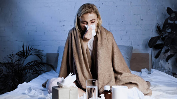 sick woman sneezing into napkin while sitting on bed near medications