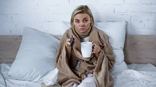 お茶を飲みながら毛布の下に座ってテレビを見ている病気の女性 — ストック写真