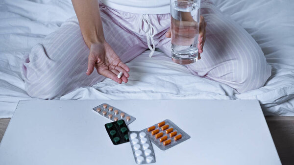 cropped view of sick woman holding pill and water near table with medications