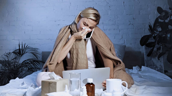 ill freelancer talking on mobile phone while sneezing into paper napkin near laptop in bedroom
