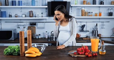 Hamile kadın mutfakta taze yemek yakınında akıllı telefondan konuşuyor. 