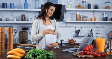 Gülümseyen hamile kadın mutfaktaki taze yemeğin yanında laptopta görüntülü sohbet ediyor. 