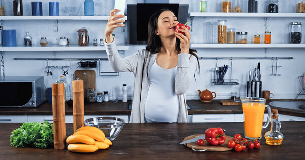 Pregnant woman smelling bell pepper and taking selfie on smartphone in kitchen 