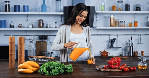 Mujer Embarazada Sonriente Vertiendo Jugo Naranja Cerca Verduras Orgánicas Cocina — Foto de Stock