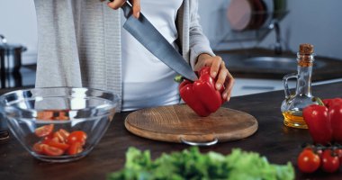 Sebzelerin yanında bıçak ve dolma biber tutan bir kadın ön planda bulanık görünüyor. 
