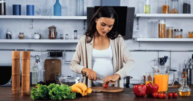 Hamile kadın mutfak masasındaki taze malzemelerin yanında kiraz domatesi kesiyor. 