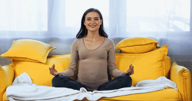 Cheerful pregnant woman sitting in lotus pose on couch  clipart