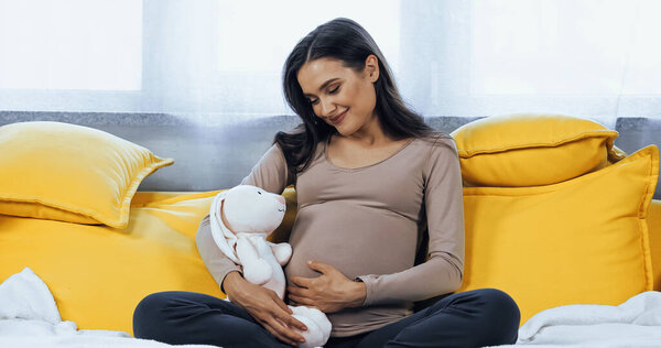 Young pregnant woman holding soft toy while sitting on couch 