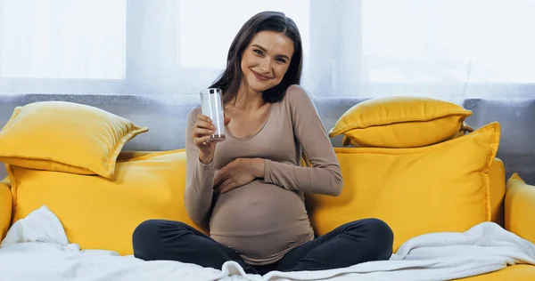 快乐的孕妇在家拿着一杯黄色沙发的牛奶 — 图库照片
