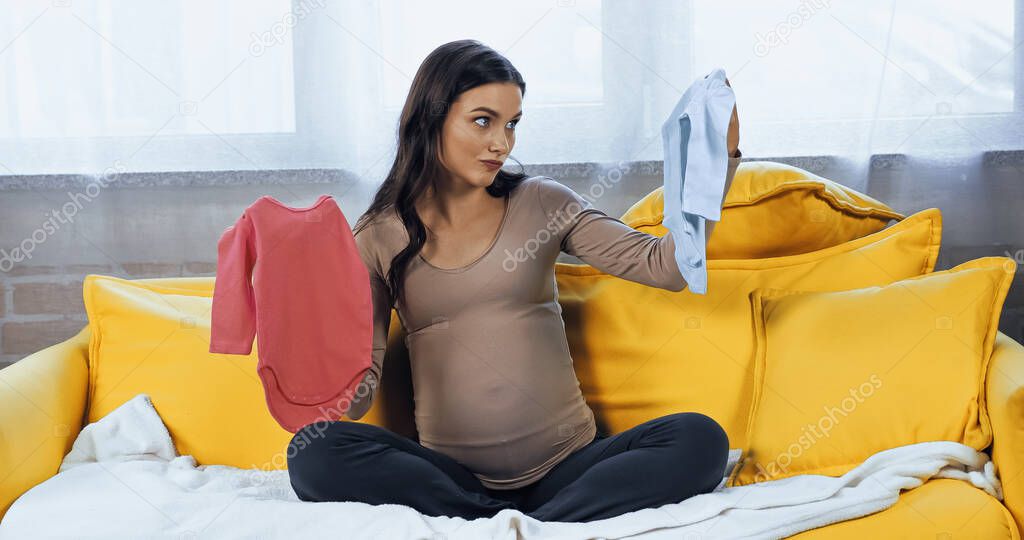 Pregnant woman looking at baby bodysuit on couch 