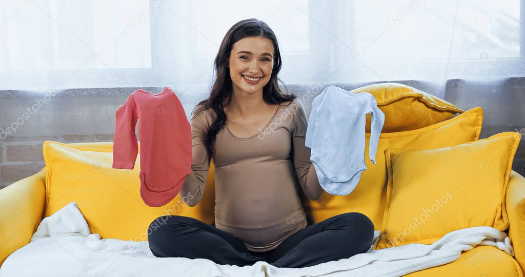 Smiling pregnant woman holding baby clothes and looking at camera 