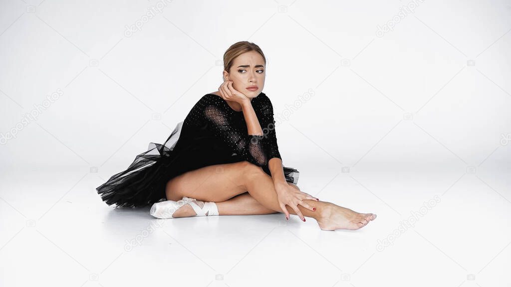 sad ballerina in tutu skirt looking away while stretching on white