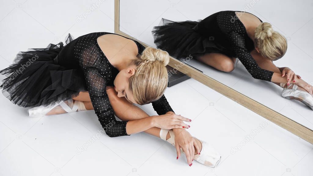 flexible ballerina in tutu skirt looking away while stretching near mirror on white