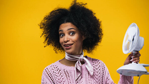 Cheerful african american woman holding electric fan isolated on yellow