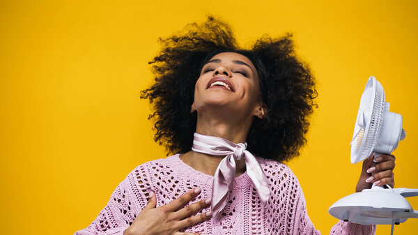 Happy african american woman holding electric fan isolated on yellow