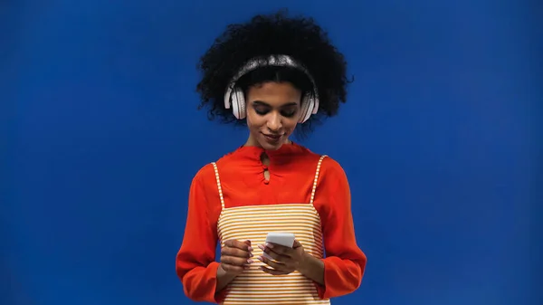 Αφροαμερικανή Γυναίκα Που Χρησιμοποιεί Ακουστικά Και Smartphone Απομονώνονται Μπλε — Φωτογραφία Αρχείου