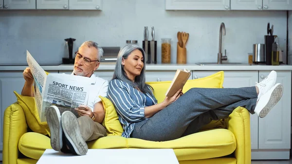 上了年纪的跨种族夫妇在家里的沙发上看书和看报纸 — 图库照片